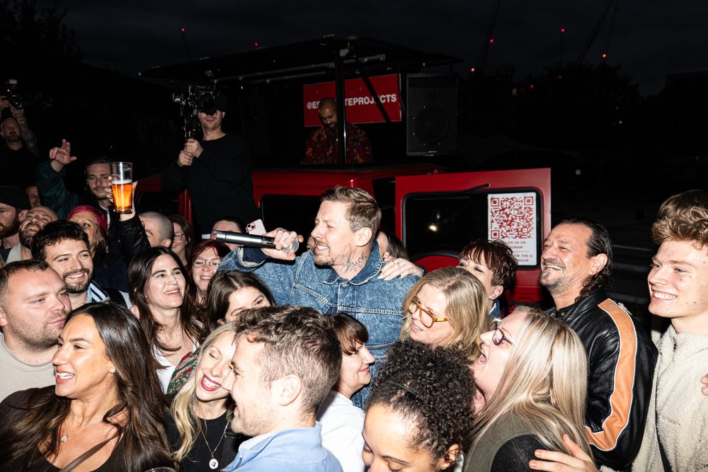 Professor Green singing among a crowd of fans in front of the red stage vehicle at night